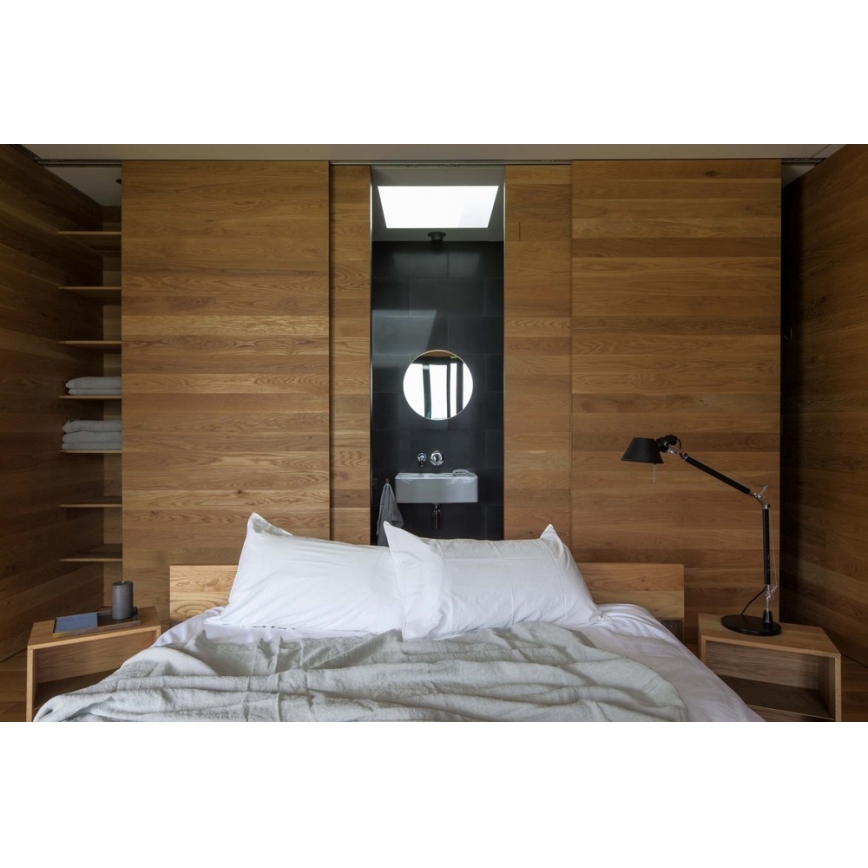 Drevená posteľ s bielou posteľnou bielizňou a svetlosivou prikrývkou stojí v spálni. Za posteľou sú drevené posuvné dvere s otvorenou časťou, odhaľujúcou malú kúpeľňu s umývadlom a okrúhlym zrkadlom. Na nočných stolíkoch sú stolové lampy.