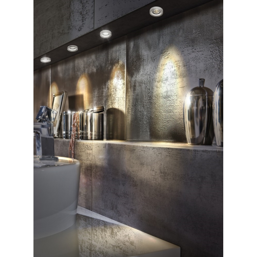 Svietidlá osvetľujú kúpeľňu. Svietidlá sú zabudované do steny nad umývadlom a poličkou s doplnkami. Stena a polička sú tmavosivé.