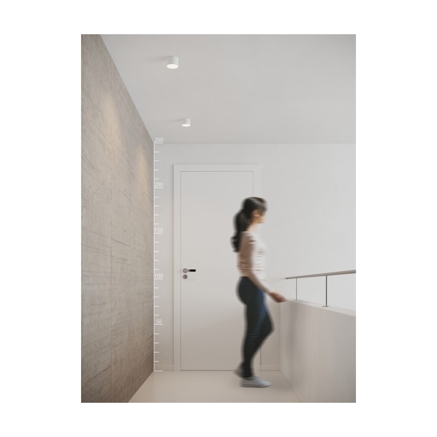 Žena kráča chodbou s betónovou stenou a bielymi dverami. Na strope sú tri zapustené bodové svetlá. Na stene je umiestnená mierka v centimetroch (50, 100, 150, 200, 250).