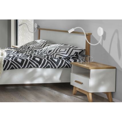 Dve nástenné lampy s flexibilným ramenom osvetľujú posteľ. Posteľ s čierno-bielou prikrývkou stojí v spálni pri nočnom stolíku.