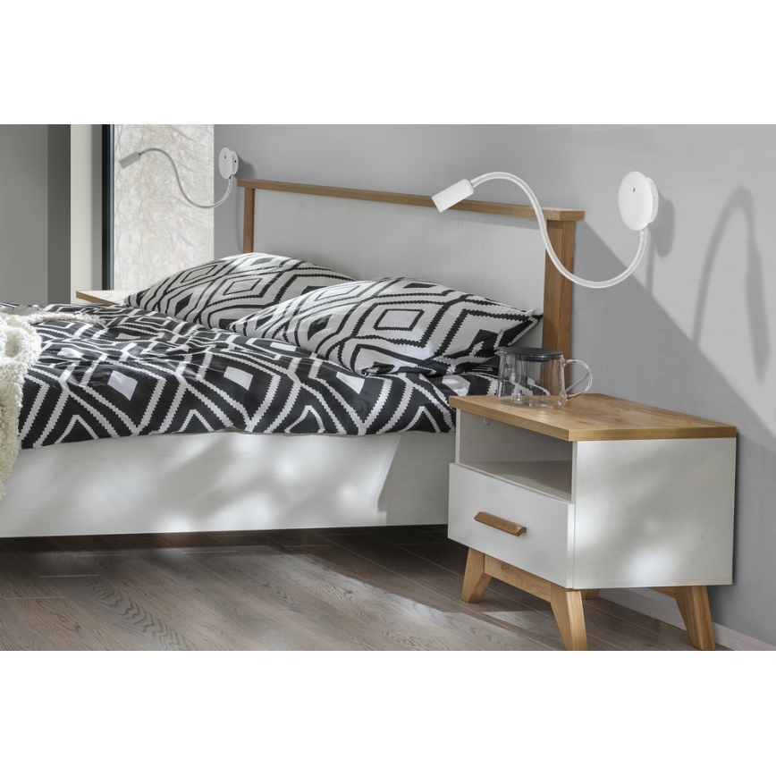 Dve nástenné lampy s flexibilným ramenom osvetľujú posteľ. Posteľ s čierno-bielou prikrývkou stojí v spálni pri nočnom stolíku.