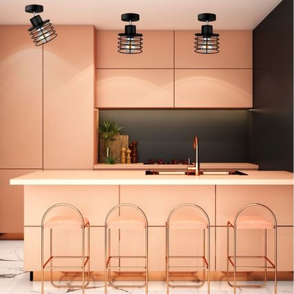 Tri stropné svietidlá s kovovými tienidlami visia v kuchyni. Svietidlá sú umiestnené nad kuchynským ostrovčekom s ružovými stoličkami.