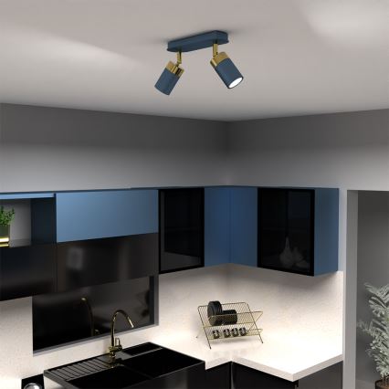 Dvojité nástenné svietidlo svieti v kuchyni. Svietidlo je tmavomodré s mosadznými detailmi. Kuchyňa má tmavomodré a čierne skrinky a svetlú pracovnú dosku.
