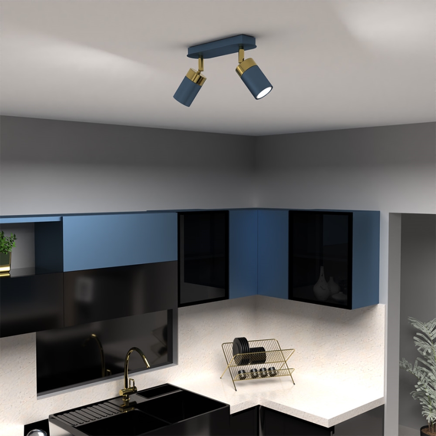 Dvojité nástenné svietidlo svieti v kuchyni. Svietidlo je tmavomodré s mosadznými detailmi. Kuchyňa má tmavomodré a čierne skrinky a svetlú pracovnú dosku.