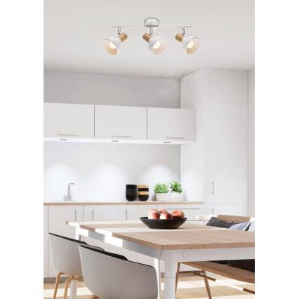 Trojramenná stropná lampa s bielymi tienidlami a drevenými detailmi je osvetlením v kuchyni. Lampa je pripevnená k stropu nad jedálenským stolom. V pozadí je svetlá kuchynská linka a jedálenská súprava.