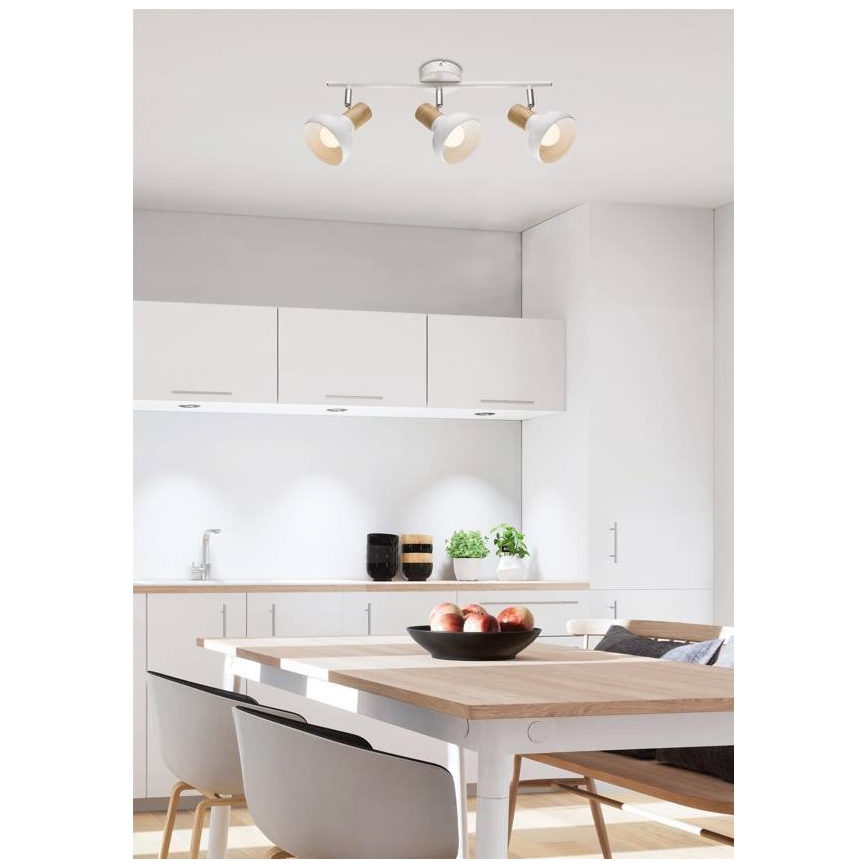 Trojramenná stropná lampa s bielymi tienidlami a drevenými detailmi je osvetlením v kuchyni. Lampa je pripevnená k stropu nad jedálenským stolom. V pozadí je svetlá kuchynská linka a jedálenská súprava.
