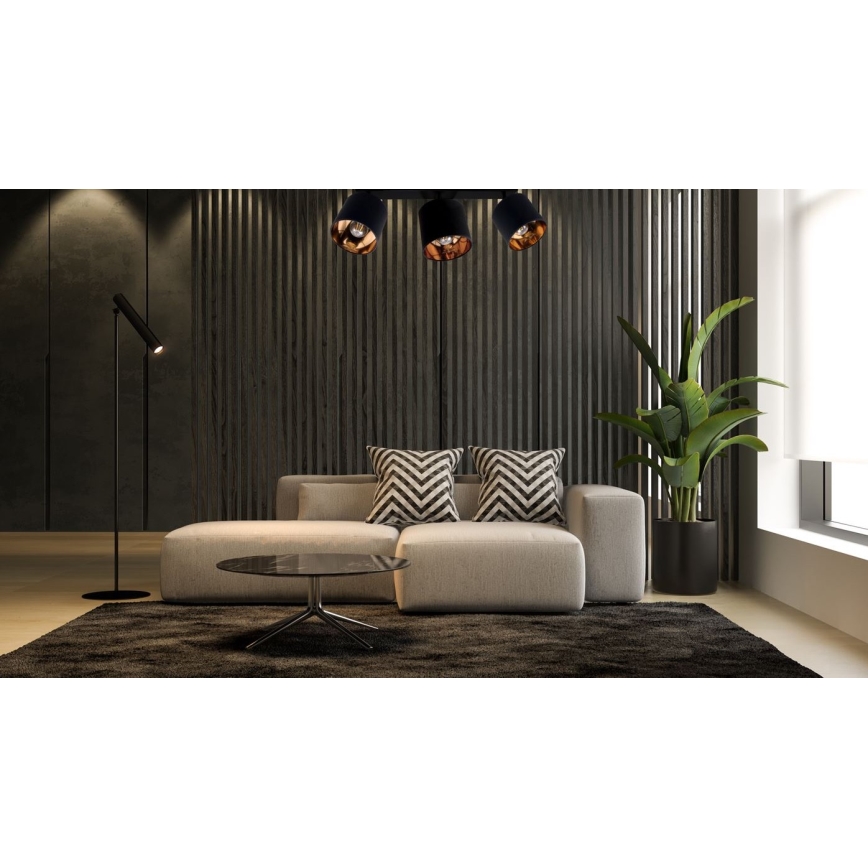 Trojité stropné svietidlo s čiernymi tienidlami a medenými detailmi osvetľuje modernú sedačku s vankúšmi v obývacej izbe s tmavohnedou stenou a rastlinou v črepníku. Na podlahe je tmavý koberec a stolík.