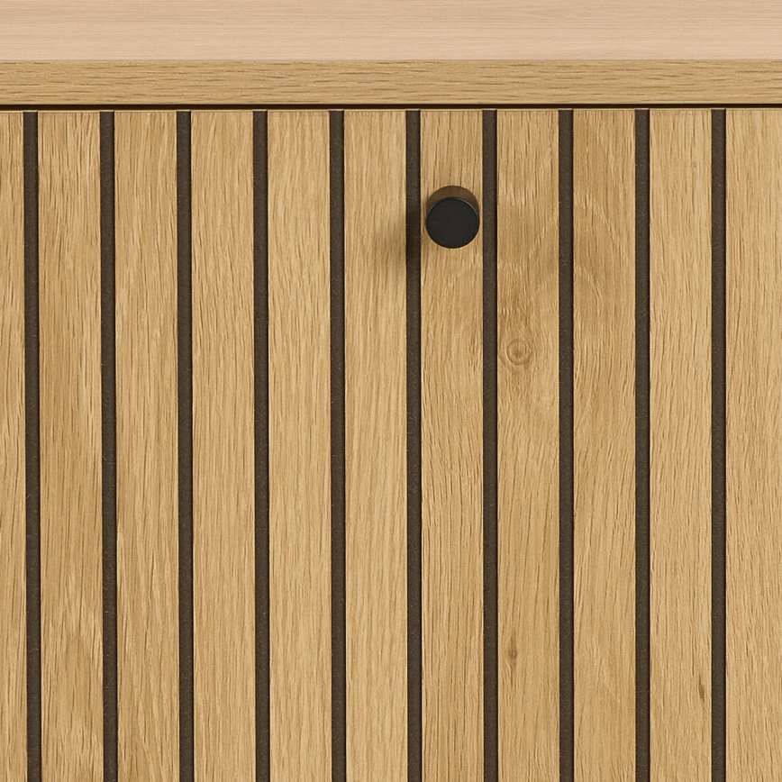 Drevené dvierka s vertikálnymi drážkami a čiernou kľučkou sú umiestnené na svetlom drevenom nábytku.