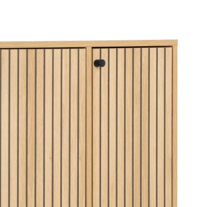 Drevená skrinka s dvoma dvierkami, zdobenými vertikálnymi drážkami, je zobrazená zblízka. Dvierka sú opatrené malými čiernymi úchytkami. Skrinka je svetlohnedá.