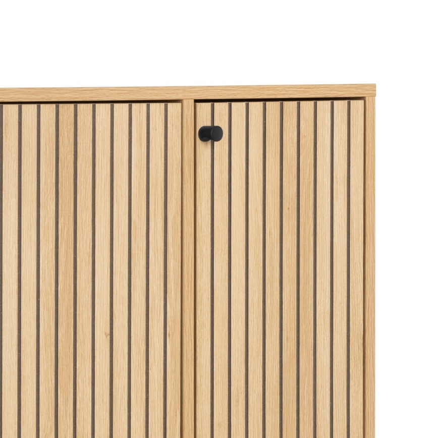 Drevená skrinka s dvoma dvierkami, zdobenými vertikálnymi drážkami, je zobrazená zblízka. Dvierka sú opatrené malými čiernymi úchytkami. Skrinka je svetlohnedá.