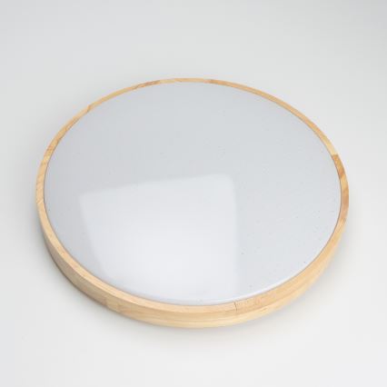 Svetlo — je umiestnené — na bielom pozadí. Má okrúhly tvar, s bielym difúzorom a dreveným rámom.