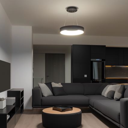 Svietidlo s kruhovým tienidlom visí zo stropu. Rohová sedačka s vankúšmi sa nachádza v tmavo zariadenej obývacej izbe. V pozadí je tmavá kuchyňa.