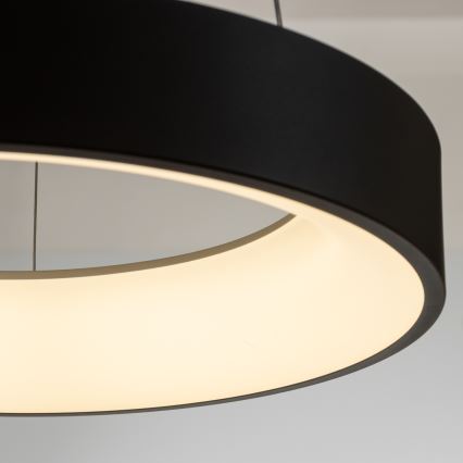 Kruhová lampa visí zo stropu. Svieti teplým svetlom. Lampa je čiernej farby s bielym vnútorným kruhom.