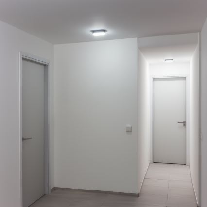 Dve biele dvere sa nachádzajú v úzkej, svetlej chodbe s dlaždicovou podlahou a stropnými svetlami. Jedny dvere sú vľavo, druhé vpravo na konci chodby.