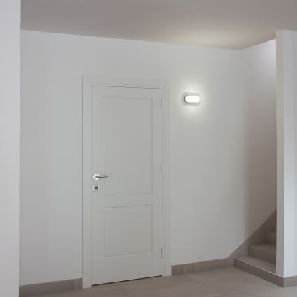 Biele dvere sú umiestnené v bielej miestnosti s dlažbou. Vedľa dverí je zapnutá nástenná lampa. Vpravo sú schody.