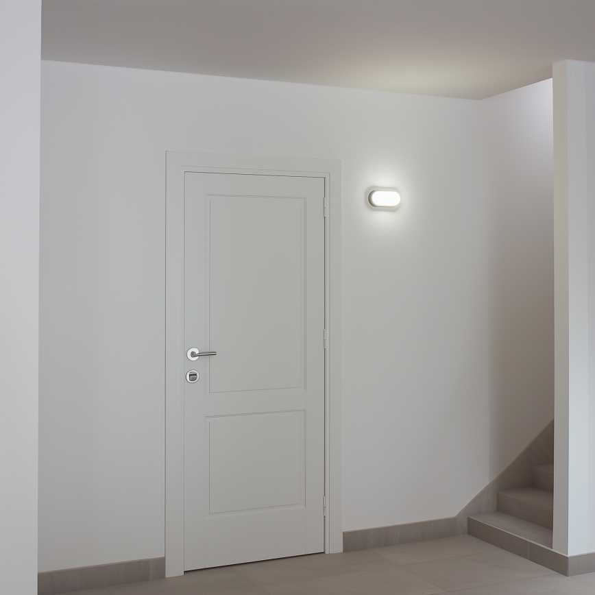 Biele dvere sú umiestnené v bielej miestnosti s dlažbou. Vedľa dverí je zapnutá nástenná lampa. Vpravo sú schody.