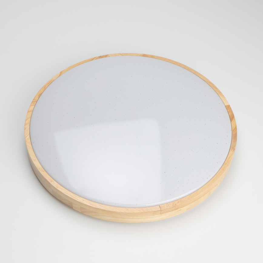 Svetlo — je umiestnené — na bielom pozadí. Má okrúhly tvar, s bielym difúzorom a dreveným rámom.