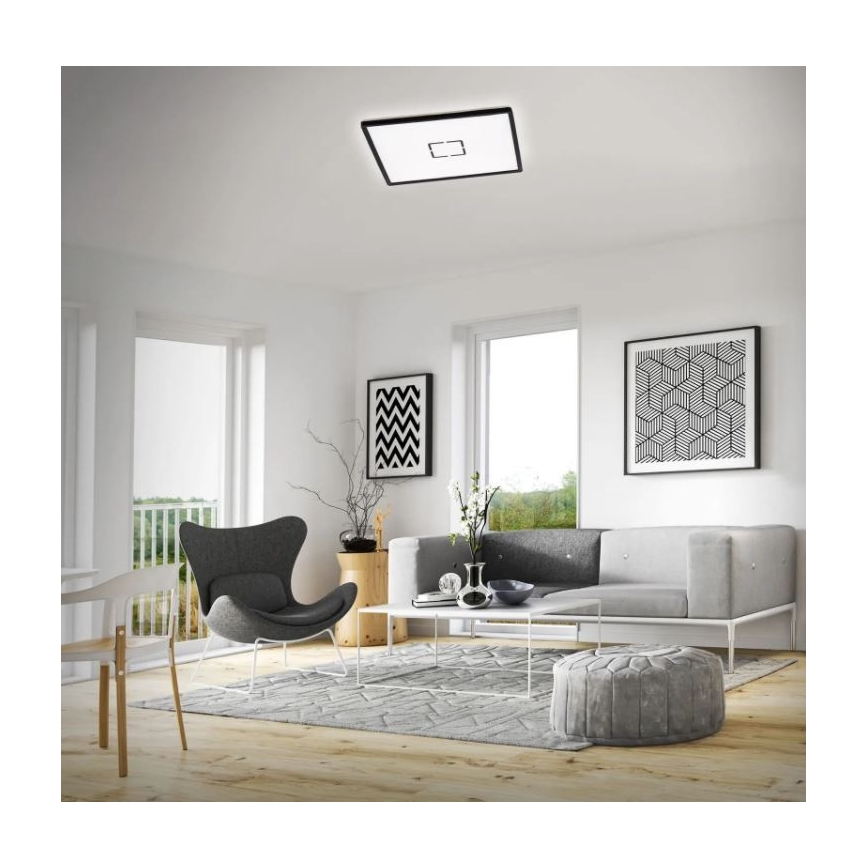 Stropné svietidlo svieti v moderne zariadenej obývacej izbe. Svietidlo je štvorcové, čiernej farby s bielym svetlom. Izba obsahuje sedaciu súpravu, kreslo a konferenčný stolík.