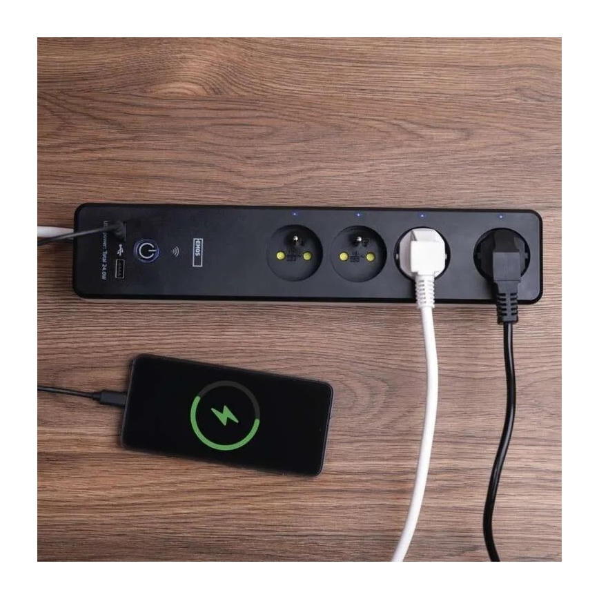 Predlžovací kábel s viacerými zásuvkami a USB portom je zapojený do elektrickej siete a nabíja mobilný telefón. Na drevenom stole sú pripojené rôzne káble a zariadenia. Na predlžovacom kábli je nápis „Unipower Total 24/24W“.