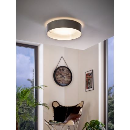 Stropné svietidlo svieti v miestnosti. Svietidlo je okrúhle, sivé s bielym vnútorným difúzorom. V miestnosti je vidieť kreslo, hodiny a rastlinu.