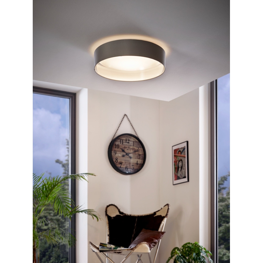 Stropné svietidlo svieti v miestnosti. Svietidlo je okrúhle, sivé s bielym vnútorným difúzorom. V miestnosti je vidieť kreslo, hodiny a rastlinu.