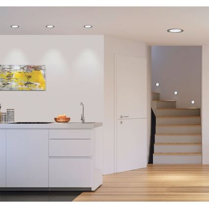 Moderná kuchynská linka s integrovaným drezom je osvetlená vstavanými bodovými svetlami. Vedľa sa nachádza schodisko s podobným osvetlením. V pozadí je obraz a svetlé drevené podlahy.