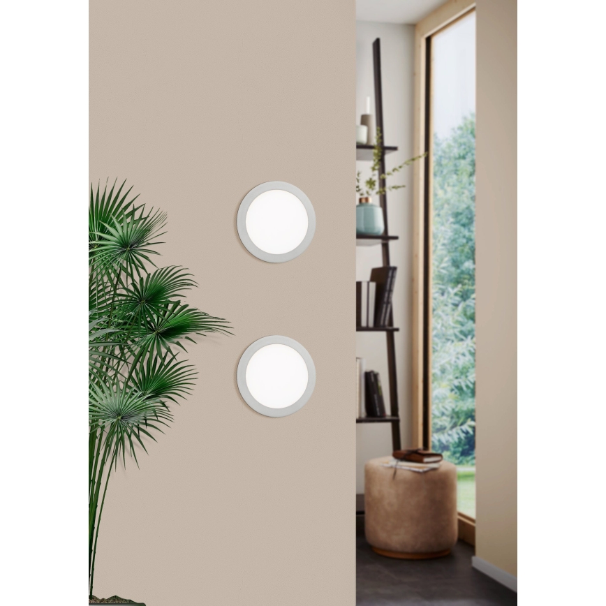 Dve okrúhle nástenné svietidlá sú umiestnené na svetlohnedej stene. Svietidlá sú osvetlené. V pozadí je vidieť izbu s rastlinou, knižnicou a oknom.