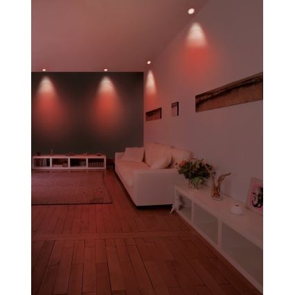 Svetlá svietia v červenom odtieni v obývacej izbe. Biela sedačka je umiestnená pred bielou poličkou. Drevená podlaha a stena v tmavosivej farbe tvoria pozadie.