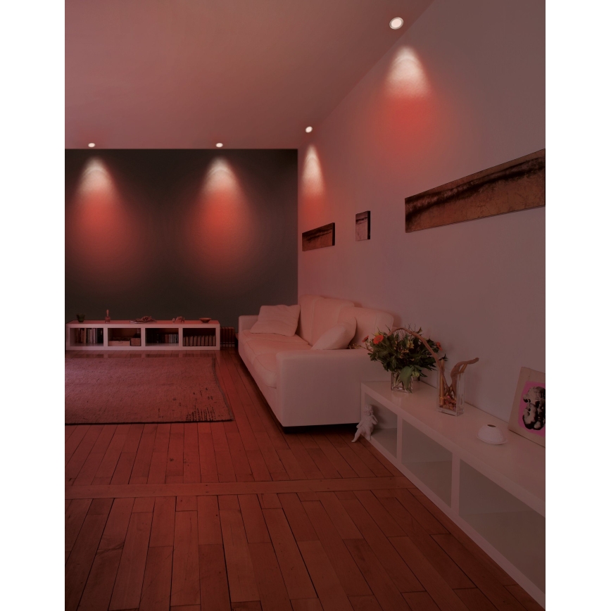 Svetlá svietia v červenom odtieni v obývacej izbe. Biela sedačka je umiestnená pred bielou poličkou. Drevená podlaha a stena v tmavosivej farbe tvoria pozadie.