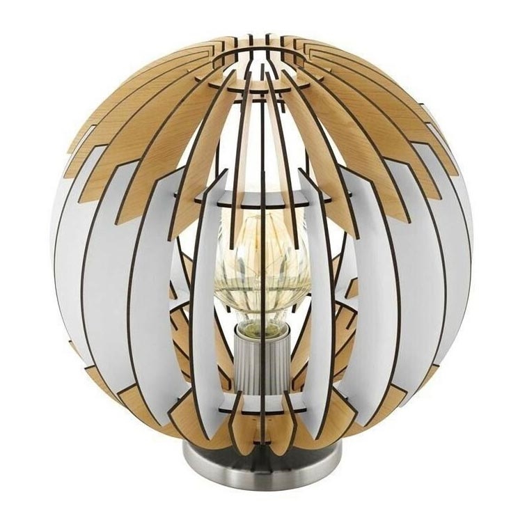 Stolová lampa s guľovou tienidlom z dreva a kovu svieti. Tienidlo je zložené z prepletených drevených lamiel v bielej a svetlohnedej farbe. Lampa stojí na kovovej podstavci.
