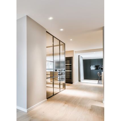 Sklenená priečka s čiernym rámom oddeľuje priestory. Priečka odráža kuchynskú linku a vinotéku. Svetlá podlaha a svetlé steny tvoria pozadí.