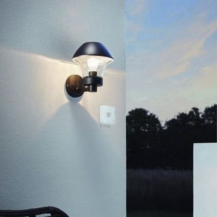 Vonkajšie nástenné svietidlo svieti na svetlosivé stene. Vedľa neho je pripevnený malý biely snímač. Pozadie tvorí zeleň stromov a večerná obloha.