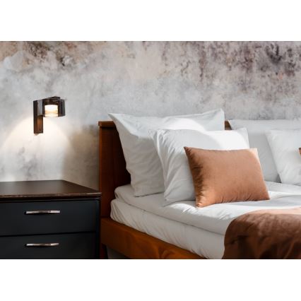 Nástená lampa svieti na posteľ s bielou posteľnou bielizňou a hnedým vankúšom. Lampa je umiestnená na stene vedľa tmavého nočného stolíka.