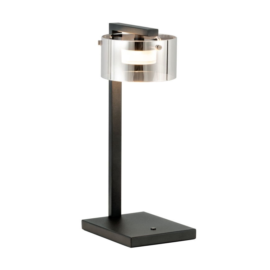 Stolová lampa svieti. Má čierny kovový stojan a priehľadný valcový tienidlo z dymového skla. Je umiestnená na bielom pozadí.
