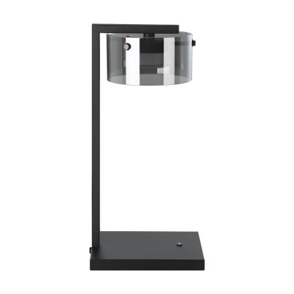 Stolová lampa s čiernym kovový rámom a priehľadným, tmavosivým skleneným tienidlom stojí na čiernej štvorcovej základni.
