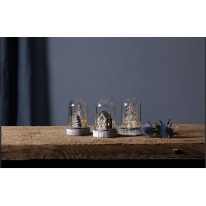 Tri sklenené dómy s drevenými vianočnými motívmi (dva stromčeky, jeden domček) s vnútorným osvetlením stoja na drevenom povrchu.