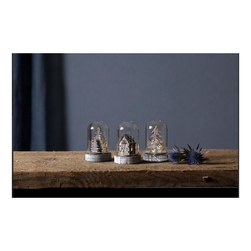 Tri sklenené dómy s drevenými vianočnými motívmi (dva stromčeky, jeden domček) s vnútorným osvetlením stoja na drevenom povrchu.