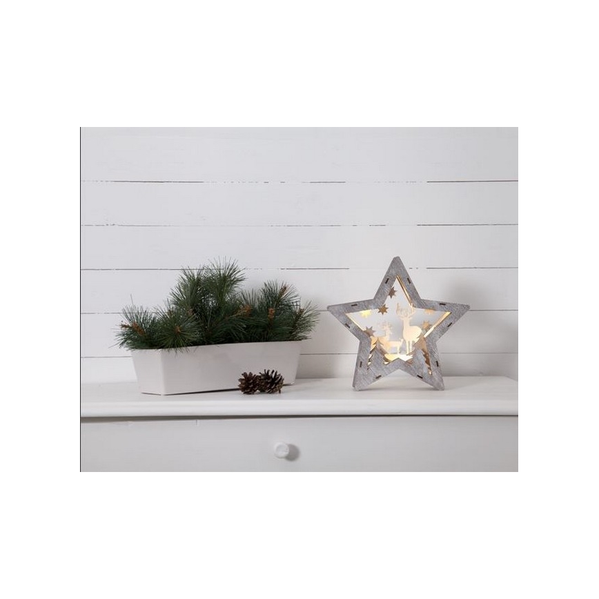 Drevená hviezda s vyrezanými jelenmi svieti. Stojí na bielom nábytku vedľa kvetináča s ihličím.