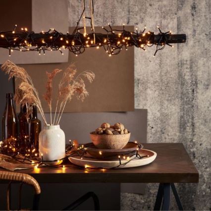 Svetelný reťaz — visí zo stropu nad stolom, kde sú rozprestreté na stole a okolo dekoratívnych predmetov. Na stole sú umiestnené váza s pampasovou trávou, miska s orechmi a drevený podnos. Pozadí tvorí textúrovaná stena.