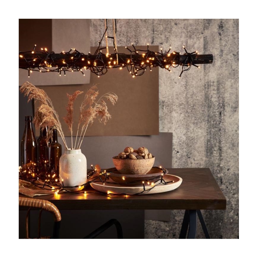 Svetelný reťaz — visí zo stropu nad stolom, kde sú rozprestreté na stole a okolo dekoratívnych predmetov. Na stole sú umiestnené váza s pampasovou trávou, miska s orechmi a drevený podnos. Pozadí tvorí textúrovaná stena.