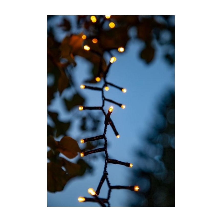 Svetlený veniec s teplými žltými LED diódami je zavesený vonku, na pozadí sú rozmazané listy a súmraková obloha.