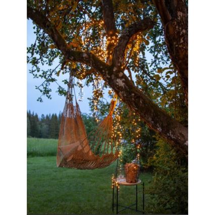 Hamaka visí na strome osvetlenom svetielkami. Vedľa je malý stolík s košíkom a pohármi. Scéna sa odohráva v záhrade pri západe slnka.