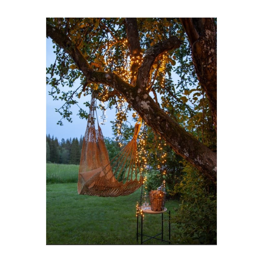 Hamaka visí na strome osvetlenom svetielkami. Vedľa je malý stolík s košíkom a pohármi. Scéna sa odohráva v záhrade pri západe slnka.