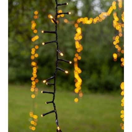 Elektrický svetelný rad s teplými žltými LED diódami visí vonku na zelenom trávniku. Svetelný rad je z čierneho kábla. V pozadí sú rozostrené ďalšie svetelné reťaze.