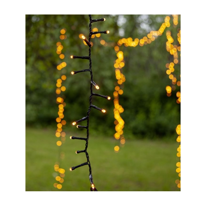 Elektrický svetelný rad s teplými žltými LED diódami visí vonku na zelenom trávniku. Svetelný rad je z čierneho kábla. V pozadí sú rozostrené ďalšie svetelné reťaze.