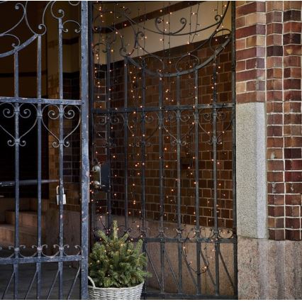 Svetielková reťaz visí pred kovovým vchodom. Reťaz je osvetlená a umiestnená na tehlovej stene. Pred vchodom sa nachádza malý ihličnatý stromček v koši.