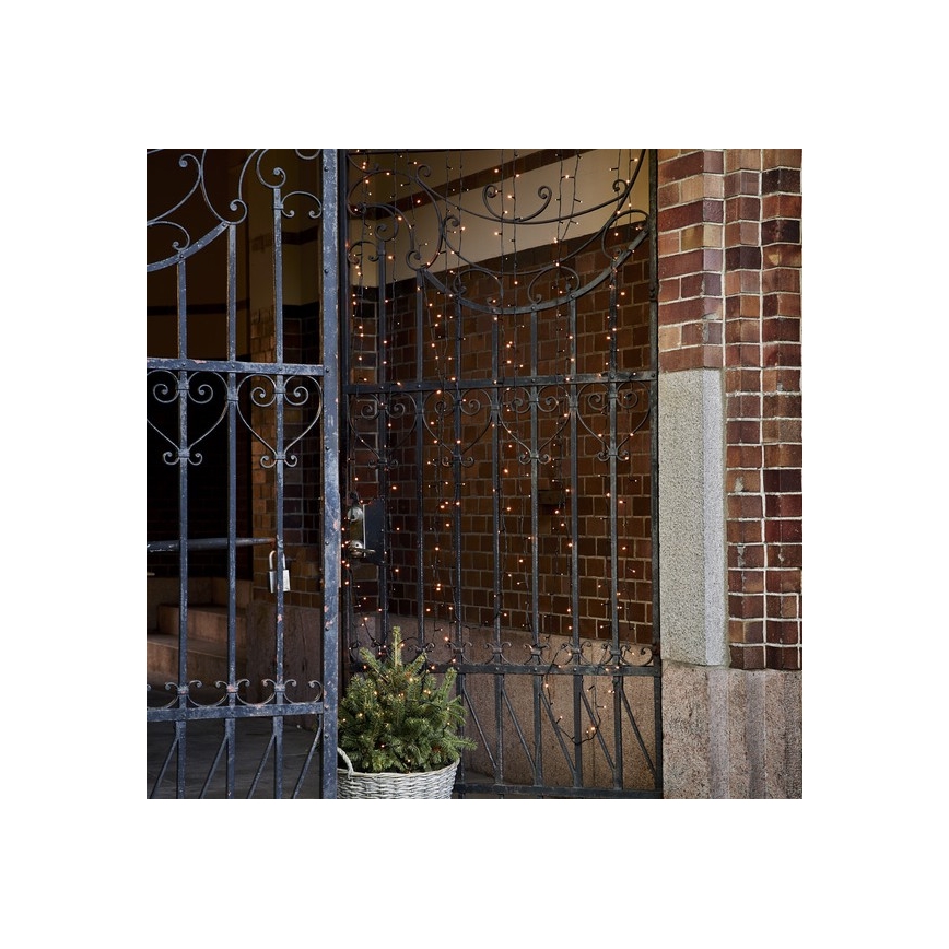 Svetielková reťaz visí pred kovovým vchodom. Reťaz je osvetlená a umiestnená na tehlovej stene. Pred vchodom sa nachádza malý ihličnatý stromček v koši.