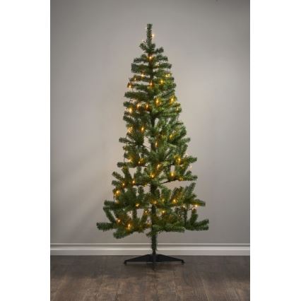 Vianočný stromček s teplými svetielkami stojí na podlahe pred svetlou stenou. Stromček je ozdobený svetelným girlandou.