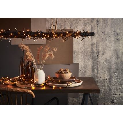 Elektrický svetelný veniec visí z drevenej tyče nad stolom. Na stole sú rozložené svetielka, váza so suchými kvetmi, miska s vlašskými orechmi a hnedé fľaše. Pozadie tvorí texturovaná stena.