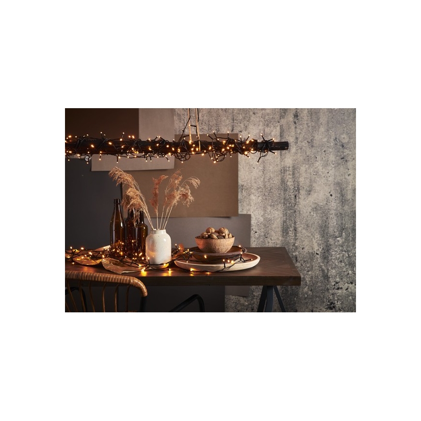 Elektrický svetelný veniec visí z drevenej tyče nad stolom. Na stole sú rozložené svetielka, váza so suchými kvetmi, miska s vlašskými orechmi a hnedé fľaše. Pozadie tvorí texturovaná stena.